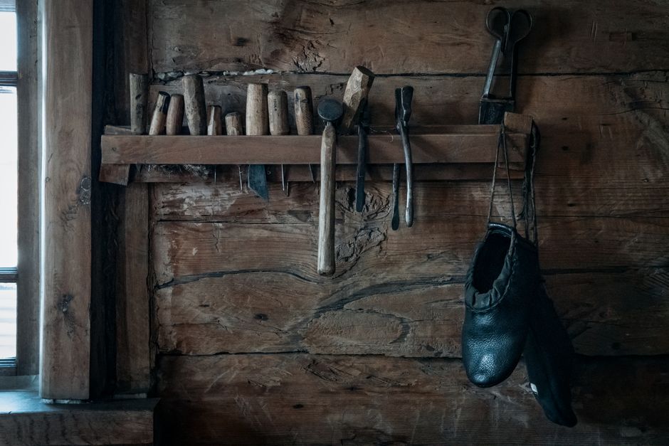 Old rustic shoes hanging on shelf on weathered shabby wooden wall near various repair tools in workshop